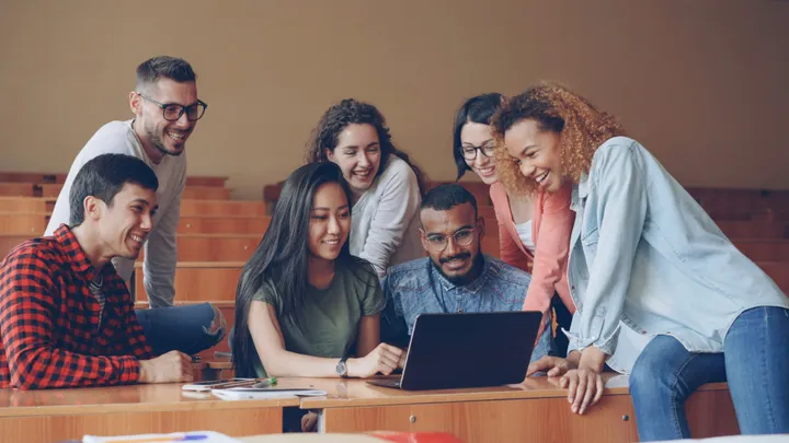 A group of students gathered around a laptop in a lecture hall, smiling and laughing together.